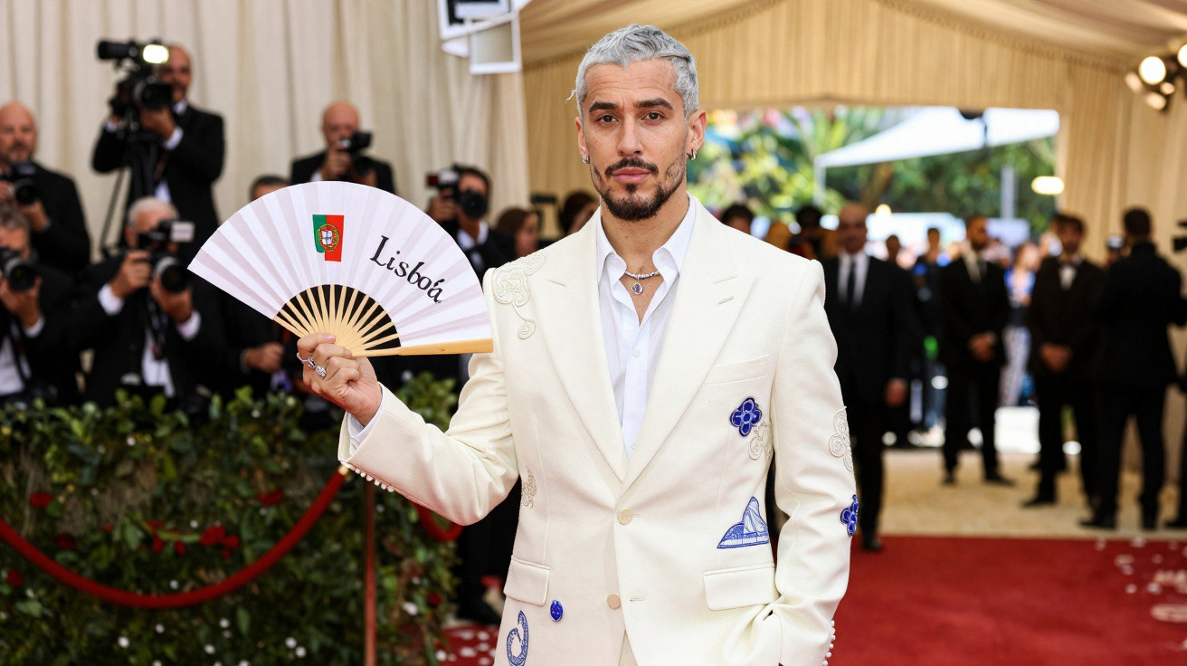 Man with grey hair in white suit holding a Lisboa fan on a red carpet with photographers in the background